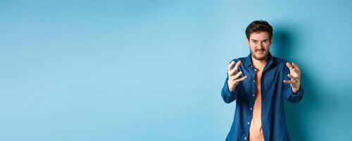 Angry tensed man wants to strangle someone, stretch out hands to choke enemy, biting lip and looking furious, standing on blue background photo