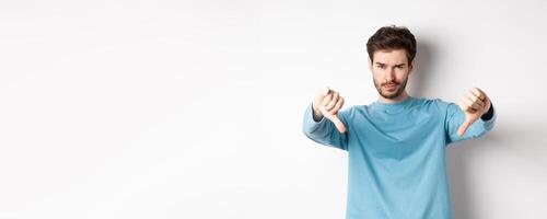 Skeptical handsome guy with beard, showing thumbs down in disapproval, dislike something bad, standing over white background photo