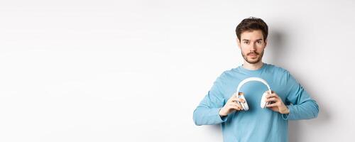 Young bearded guy in blue sweatshirt put on wireless headphones, listening music, standing on white background photo