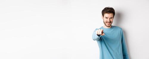 Image of confident young man smiling and pointing at camera, inviting and choosing you, beckon to join him, standing on white background photo