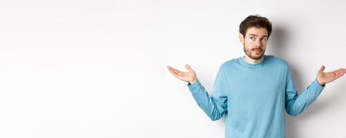 Indecisive young man with beard, shrugging shoulders and know nothing, looking away confused, standing in blue shirt over white background photo