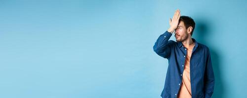 Annoyed and tired man facepalm, roll eyes and look bothered by stupid behavior, standing on blue background photo