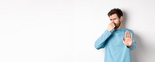 Image of caucasian man stretch out hands to stop or reject something, shut nose disgust, refusing product with bad smell, standing over white background photo