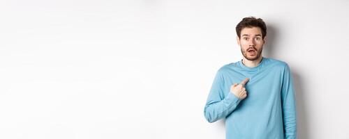 Surprised young man gasping and pointing at himself with disbelief, being chosen or named, standing over white background photo