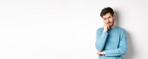 Skeptical young man listening to you with bored face, looking at camera reluctant, standing in sweatshirt over white background photo