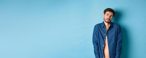 Reluctant and sad man feeling upset, looking aside at empty space with unamused face, standing on blue background photo
