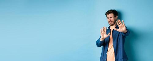 Guy cringe from something disgusting or embarrasssing, stretching out hands and asking to stop, grimacing displeased, standing on blue background photo