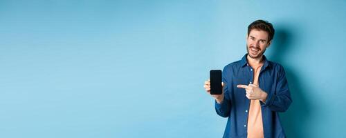 Smiling caucasian man showing on empty smartphone screen, pointing at mobile phone and looking satisfied, standing on blue background photo