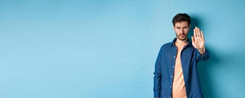 Upset man frowning and asking to stop, stretch out hand to prohibit or disagree with something bad, standing on blue background photo