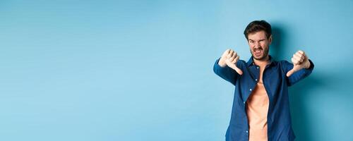 Disgusted guy express negative opinion, showing thumbs down and tongue, frowning upset, disapprove something bad, standing on blue background photo