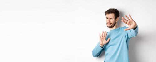 Stay away from me. Reluctant young man step back with hands stretch out in defensive gesture, protecting himself, standing over white background photo