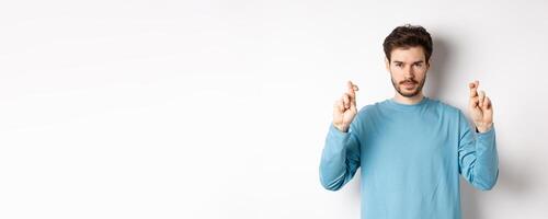 Hopeful and confident young man cross fingers for good luck, making wish or praying, anticipating results, standing over white background photo