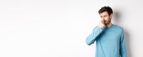 Disgusted young man in sweatshirt shut his nose, looking at something with bad smell, standing over white background photo