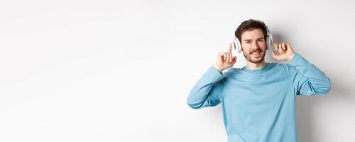 Attractive modern man with beard, smiling pleased, touching headphone on head while listening to music, standing over white background photo