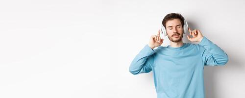 Relaxed young man enjoying favorite song, listening music in headphones with closed eyes and calm face, standing over white background photo