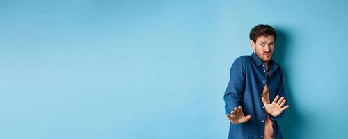 Timid young man asking to keep away, stretch out hands defensive and step back, refusing something disgusting, standing on blue background photo