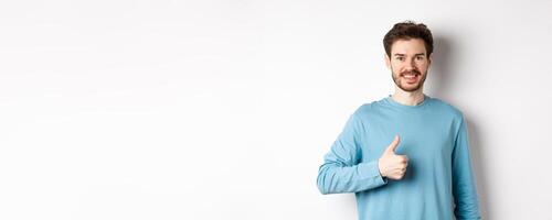 Smiling male model in casual shirt showing thumb up, approve and recommend product, praise excellent choice, standing on white background photo