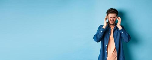 Man feeling sick, touching head and grimacing from painful migraine, having headache or feel dizzy, standing on blue background photo