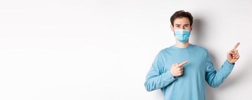 Coronavirus, health and quarantine concept. Handsome caucasian man in face mask pointing fingers right, showing advertisement, standing over white background photo