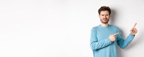 Disappointed and upset young man showing bad offer, pointing fingers right and frowning disgusted, standing with skeptical face on white background photo