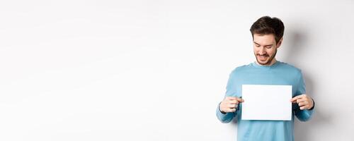 Young caucasian man looking at empty paper, reading logo, standing on white background photo