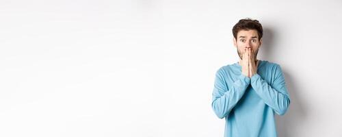 Shocked and worried young man gasping and covering mouth, look with panic at camera, standing anxious against white background photo