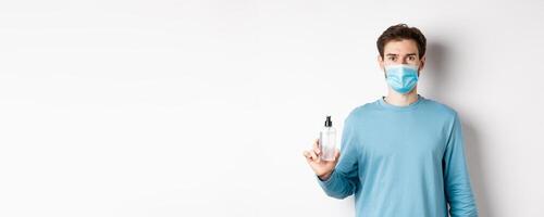 Covid-19, health and quarantine concept. Young caucasian man advice hand sanitizer, showing antiseptic and wearing medical mask, standing on white background photo