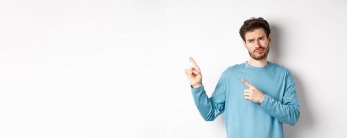 Skeptical and unimpressed young man grimacing, pointing fingers and looking at bad promotion, standing dissatisfied over white background photo