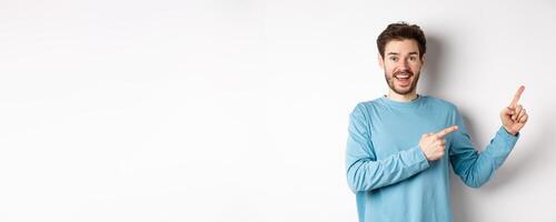 Cheerful smiling man pointing fingers right and showing banner or logo on white space for advertisements, recommending product, standing over studio background photo