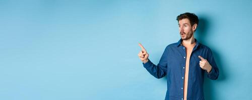 Surprised guy in casual clothes, pointing and looking left at empty space, showing advertisement, standing on blue background photo