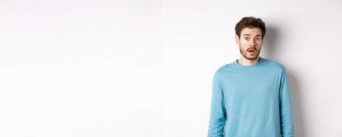 Confused caucasian guy drop jaw and stare startled at camera, standing over white background photo