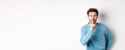 Attractive bearded man asking to keep quiet, showing taboo hush gesture and looking at camera calm, standing over white background photo