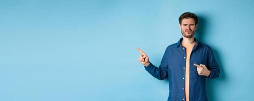 Disappointed and skeptical guy frowning, pucker lips and pointing fingers left at empty space, complaining at bad thing, standing upset on blue background photo