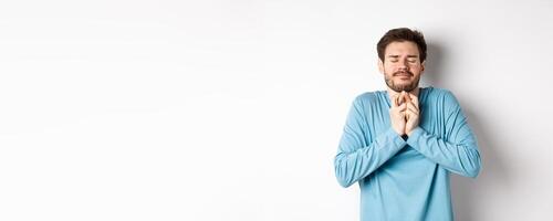 Hopeful caucasian guy in sweatshirt cross fingers for good luck. Young man making wish, praying with eyes closed and dreamy smile, standing over white background photo