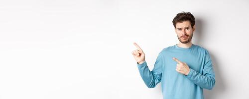 Skeptical young man with beard grimacing unsatisfied, frowning and pointing at upper left corner logo doubtful, standing upset over white background photo