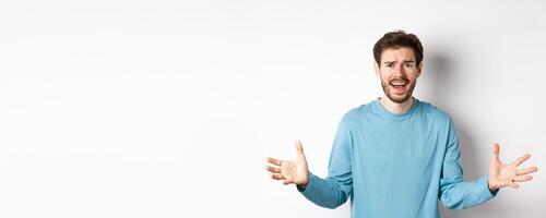 Image of frustrated and worried young man shaking hands, trying to explain something, standing anxious against white background photo