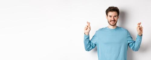 Optimistic smiling man cross fingers for good luck, making wish and waiting for results, standing over white background photo