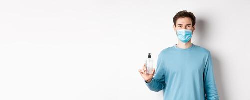 Covid-19, health and quarantine concept. Portrait of smiling man in medical mask showing hand sanitizer bottle, standing over white background photo