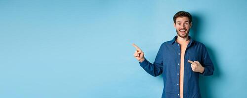 Excited and happy caucasian man smiling at camera, pointing fingers left at empty space, showing logo, standing on blue background photo