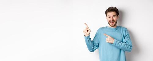 Happy caucasian man in casual blue sweatshirt, pointing fingers at upper left corner, showing link or logo on white background, smiling at camera photo