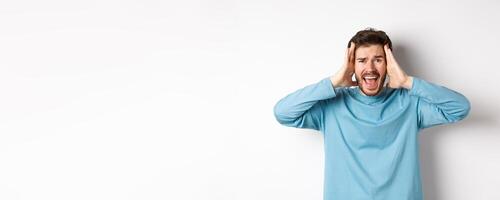 Image of young caucasian man screaming in panic, looking frustrated and shouting at camera, standing on white background photo