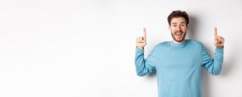 Cheerful young man showing advertisement with happy smile, pointing fingers up at awesome logo banner, standing on white background photo