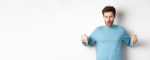 Handsome man pointing down with impressed and happy face, smiling and saying wow, standing on white background photo
