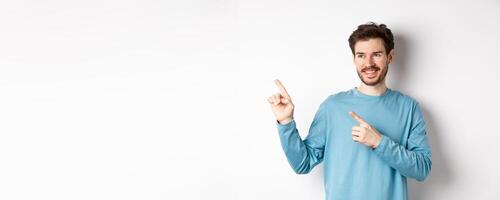 Portrait of young modern man in blue sweatshirt pointing fingers at upper left corner and looking away, smiling pleased as showing advertisement on white background photo