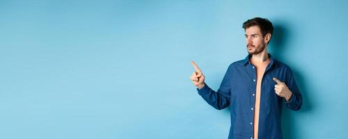 Image of young caucasian man looking and pointing aside, showing advertisement, standing on blue background photo