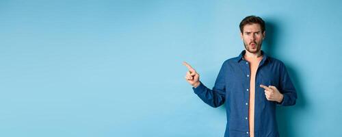 Shocked and offended young man frowning and looking hurt, pointing fingers left at empty space, complaining on something, standing on blue background photo