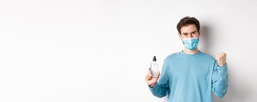 Covid-19, health and quarantine concept. Cheerful man in face mask celebrating, showing fist pump and bottle with hand sanitizer, fighting germs, white background photo