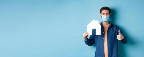 Real estate and covid concept. Young man in face mask showing paper house cutout and thumb-up, recommending agency, standing on blue background photo