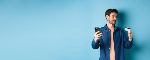 E-commerce concept. Man paying for online payment, reading credit card number and holding mobile phone, standing on blue background photo