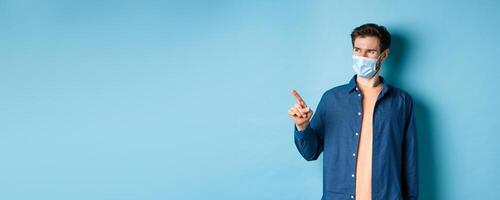 Handsome young man wearing medical mask from coronavirus, pointing finger left and looking satisfied, smiling at empty space, blue background photo